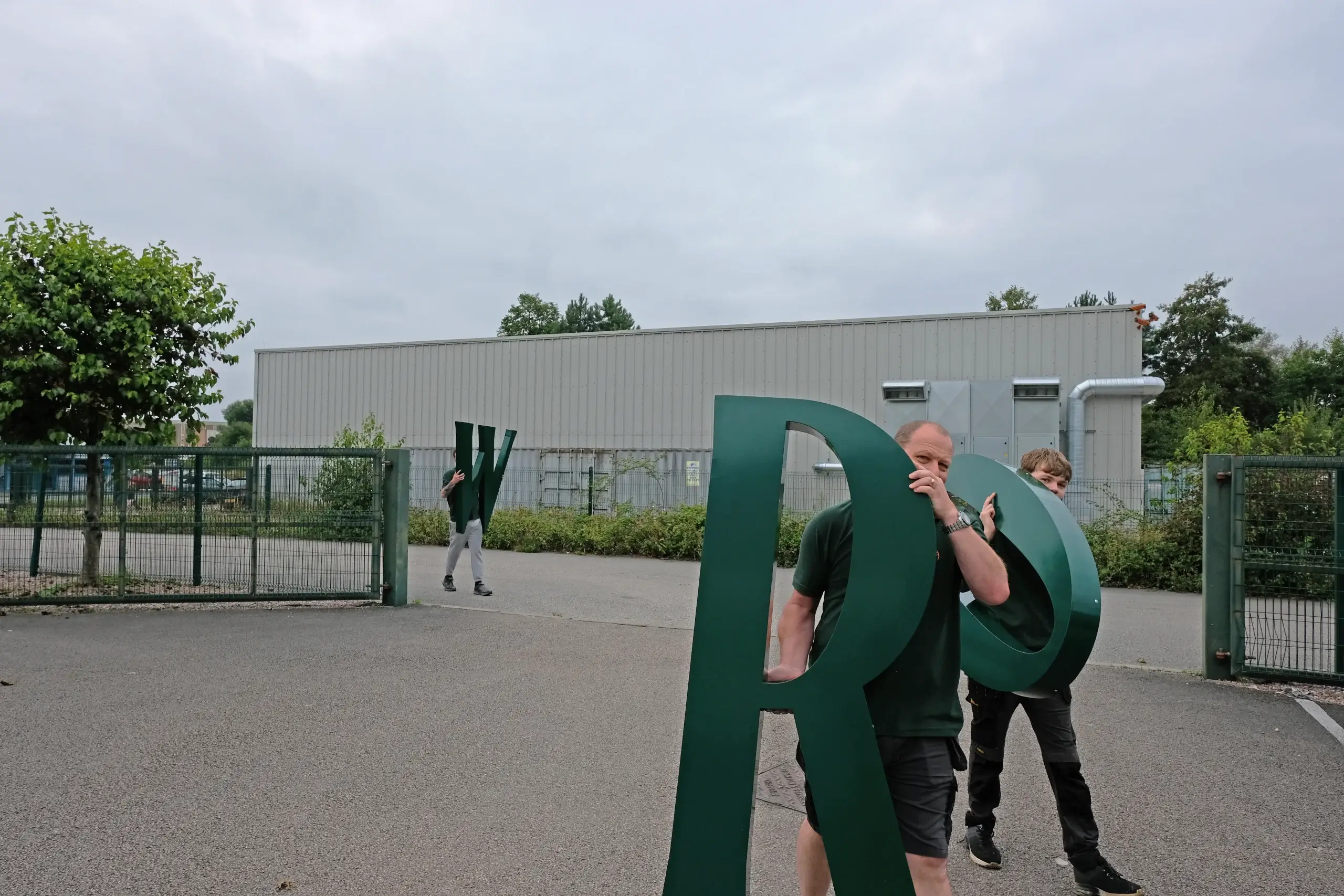 Three workers carrying large green letter sculptures (R, O, W) across an outdoor paved area near a fenced industrial building