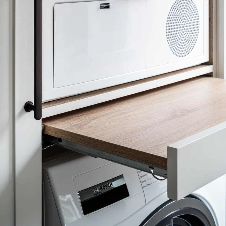 Pull-out wooden shelf above a Bosch front-load washing machine inside a white laundry cabinet with a black handle.
