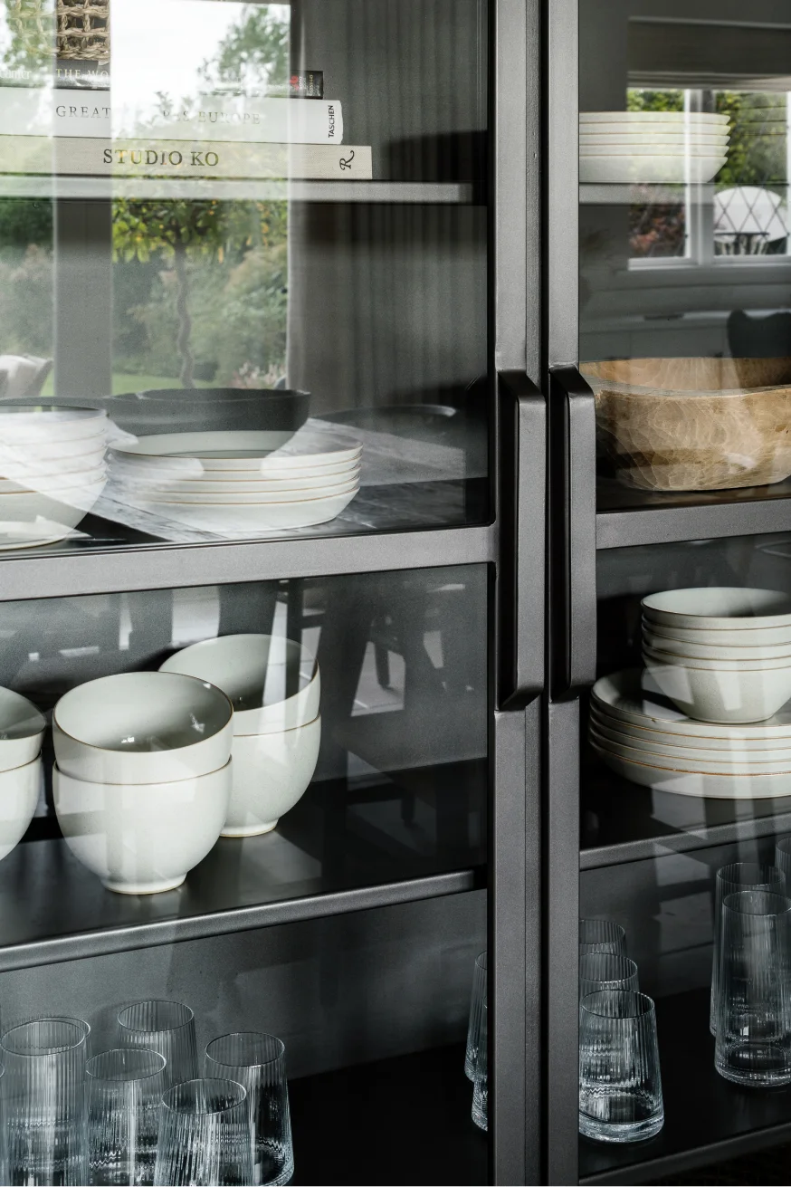 Dark metal cabinet with glass doors displaying stacked white bowls, plates, wooden bowl, and clear drinking glasses.