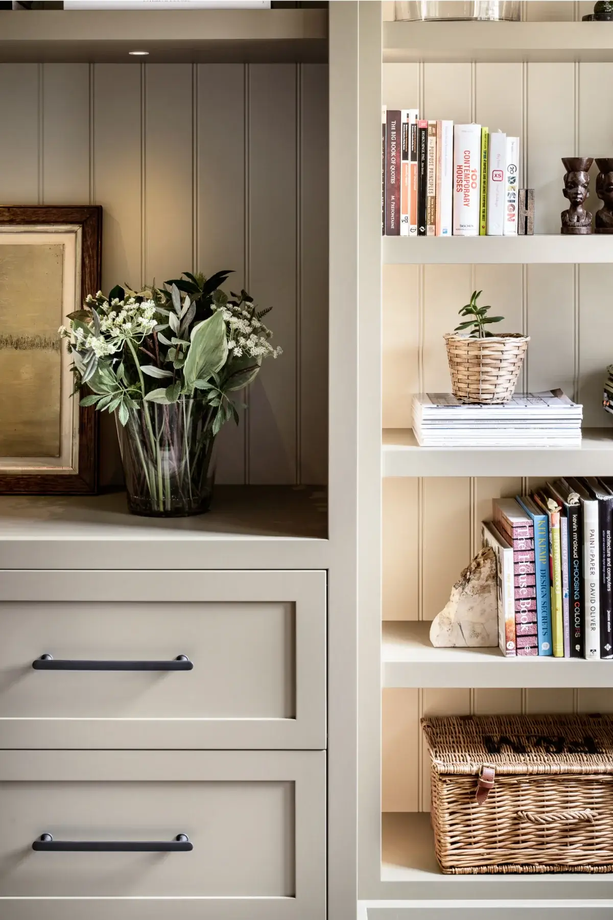 Built-in beige shelving unit with drawers, a glass vase of mixed greenery, stacked books, a small potted plant, and wicker basket.