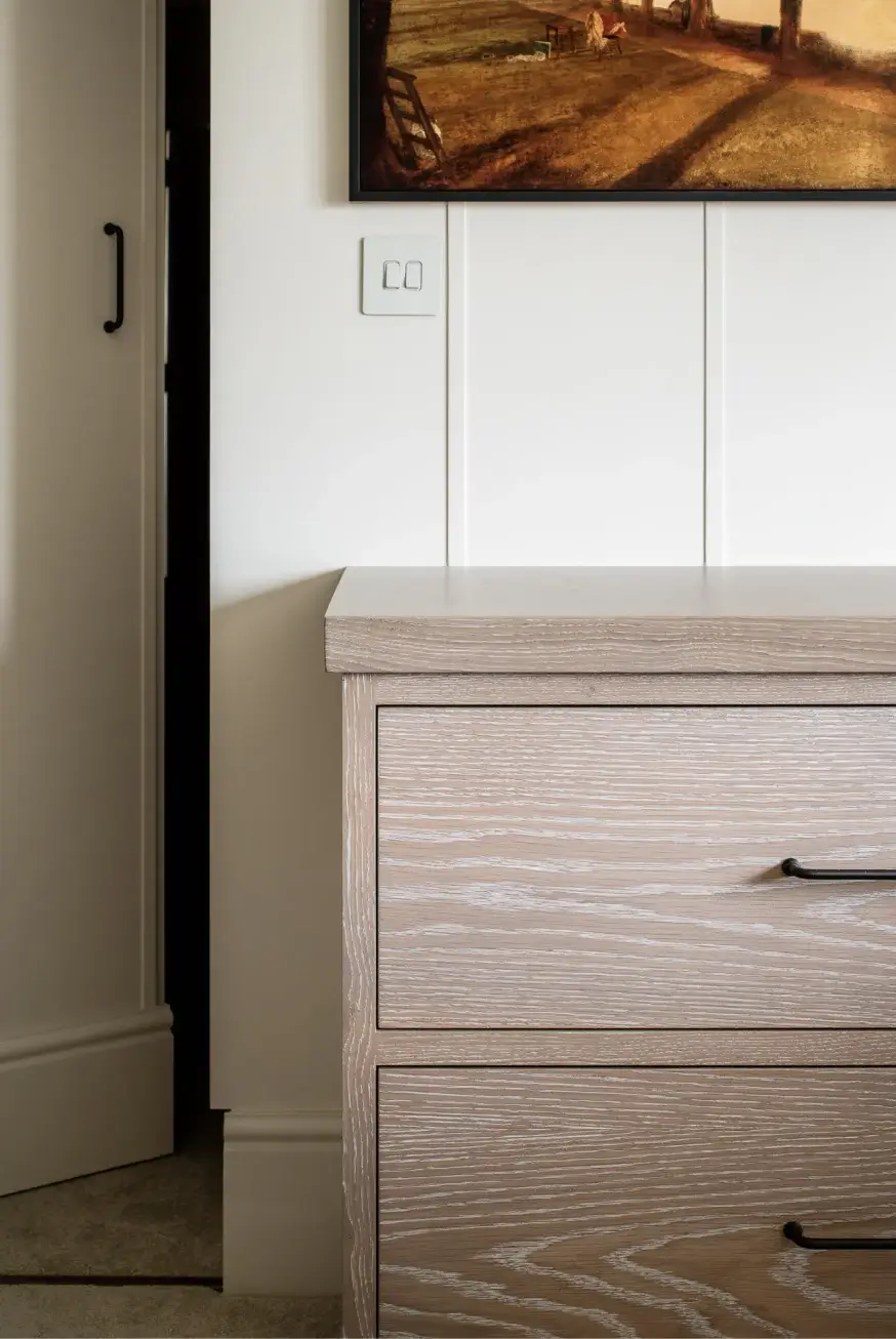 Light wood two-drawer nightstand with black metal handles against a white paneled wall and partial framed artwork above