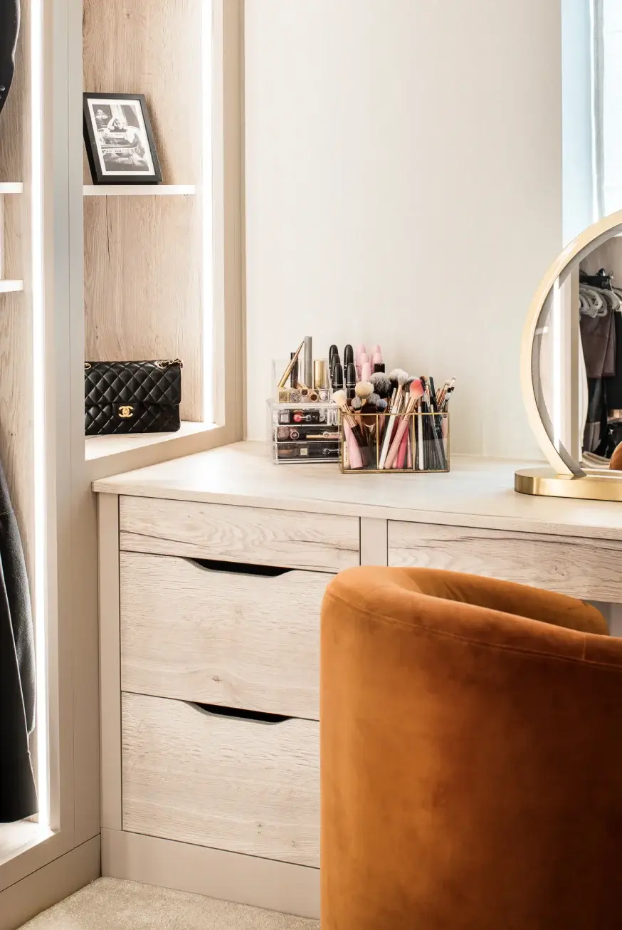 Light wood vanity with organized makeup brushes and palettes, round gold-framed mirror, quilted black purse on nearby shelf, rust velvet chair.