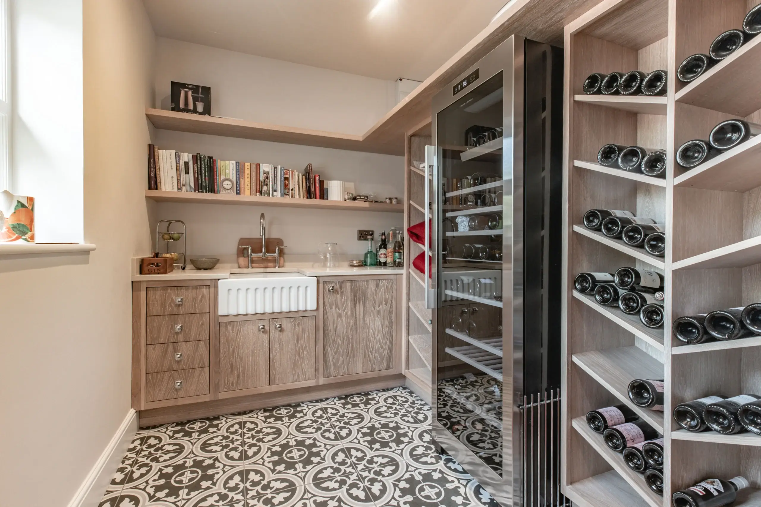Butler's pantry with wooden cabinets, farmhouse sink, open shelves of books and bottles, patterned tile floor and a glass-front wine fridge